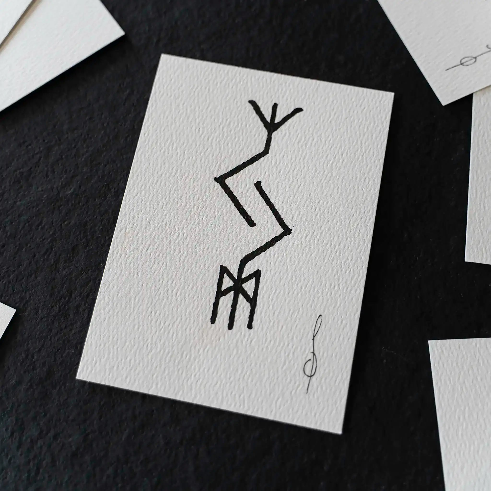 A square white art print with a black abstract line drawing, displayed on a dark textured surface with other partial prints around it.