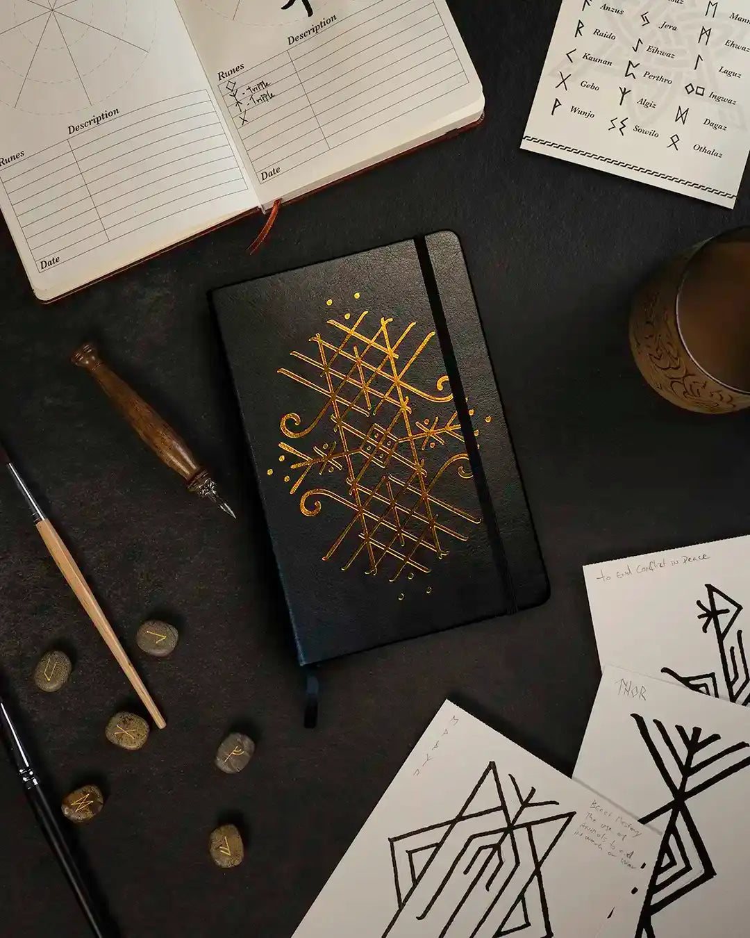 Black journal with gold Norse rune design, surrounded by rune stones, sketches, and Viking-inspired tools