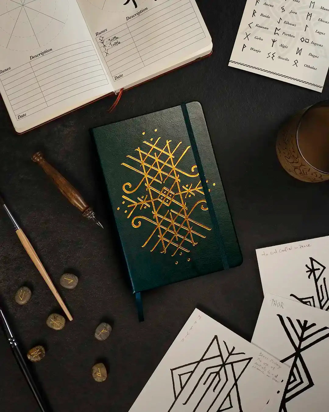 Dark green notebook with gold Norse rune design, surrounded by rune stones, sketches, and calligraphy tools
