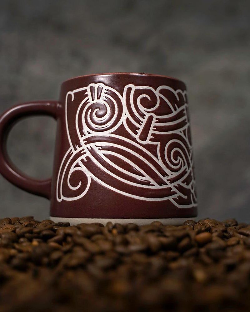 Brown ceramic mug with white Norse knotwork design, surrounded by coffee beans