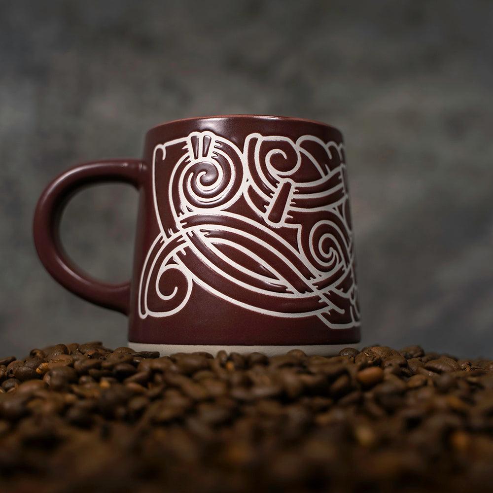 Brown ceramic mug with white Norse knotwork design, surrounded by coffee beans