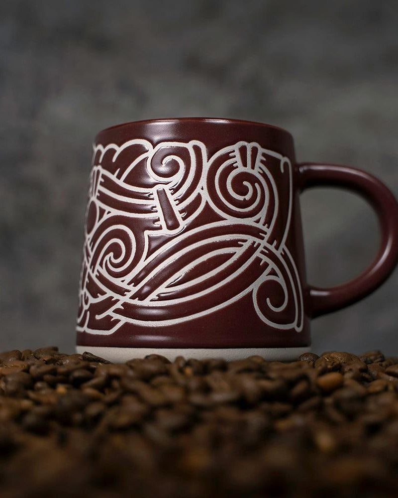 Brown ceramic mug with white Norse knotwork design, nestled in coffee beans