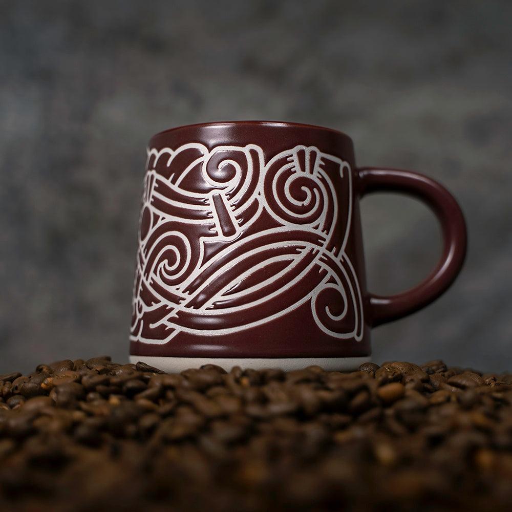 Brown ceramic mug with white Norse knotwork design, nestled in coffee beans