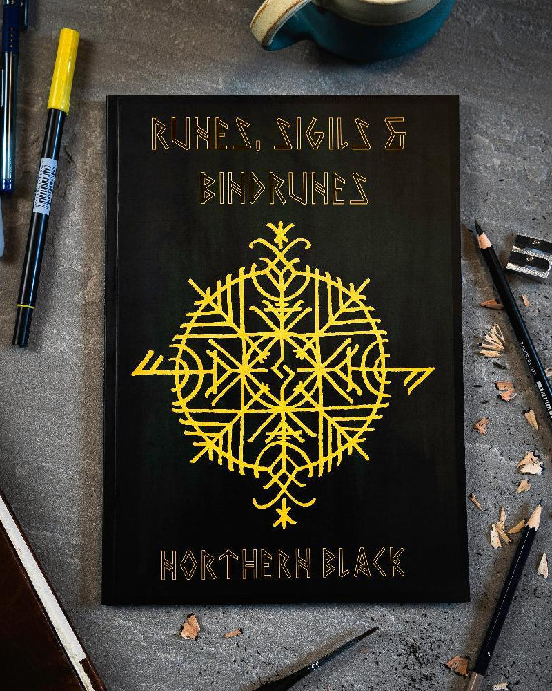 Black book with yellow Norse runes and bindrunes design, surrounded by pens and pencils