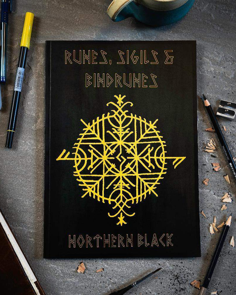 Black book with yellow Norse runes and bindrunes design, surrounded by pens and pencils