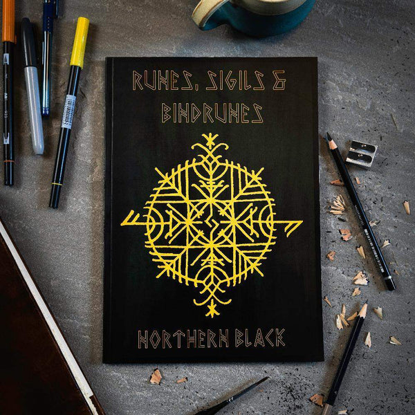 Black book with yellow Norse runes and bindrunes design, surrounded by pens and pencils