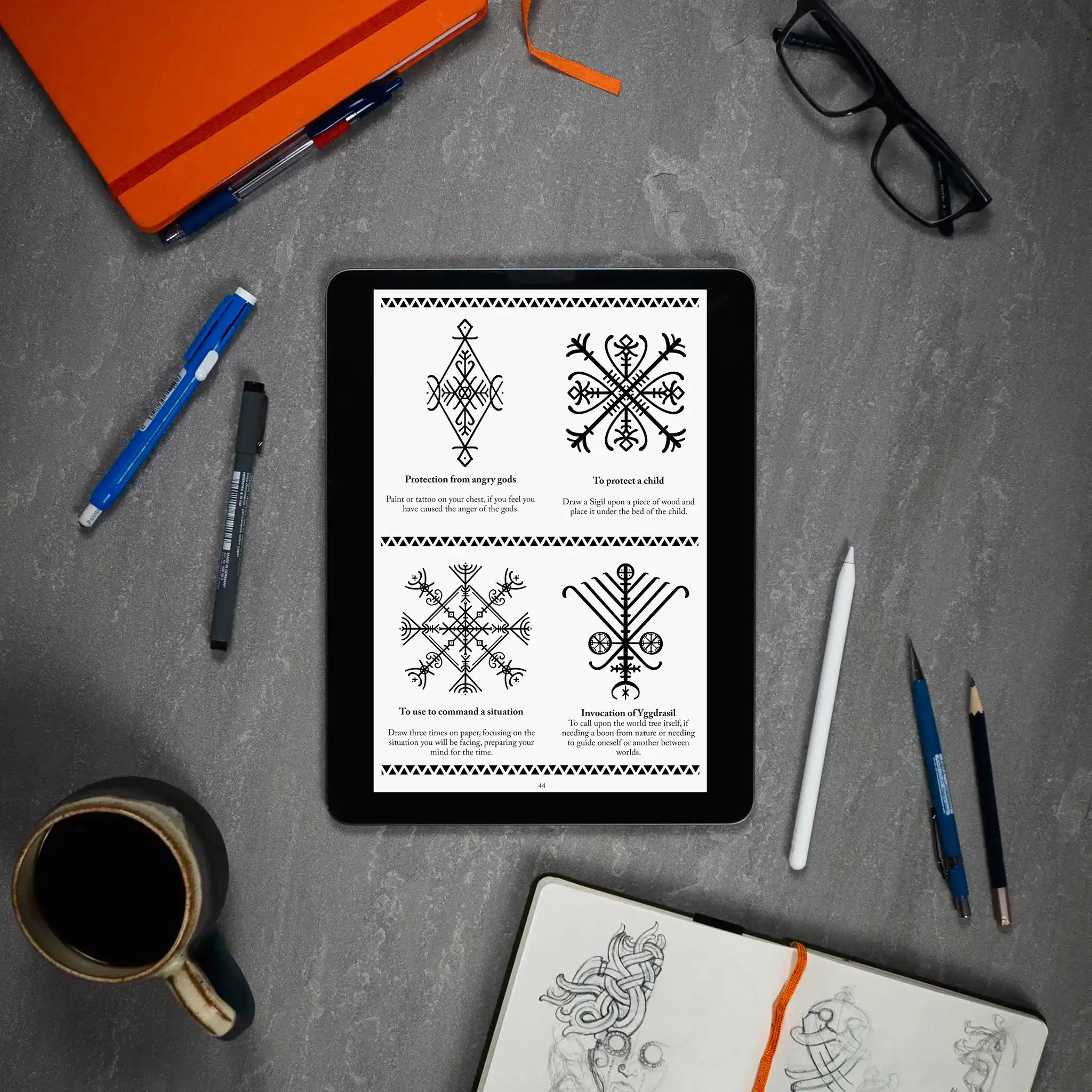 Viking rune designs and Norse symbols displayed on tablet, surrounded by sketchbook, pens, and coffee — ideal for tattoo or ritual use