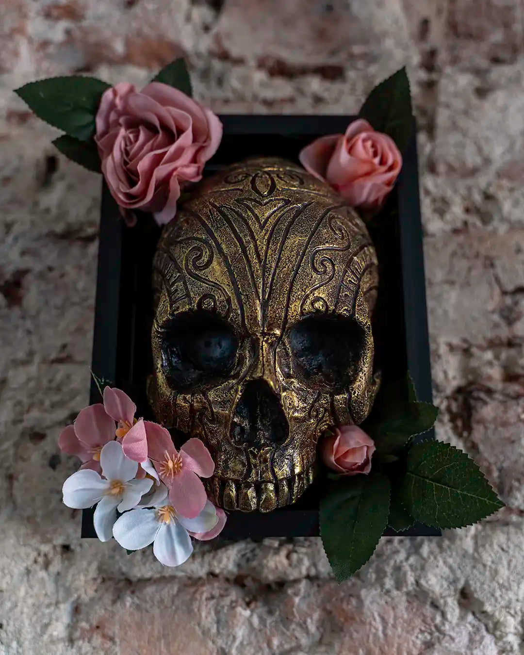 An ornate gold skull centerpiece with intricate black detailing, nestled in a black square tray and decorated with pink roses, white and pink blossoms, and green leaves against a rustic stone wall background.