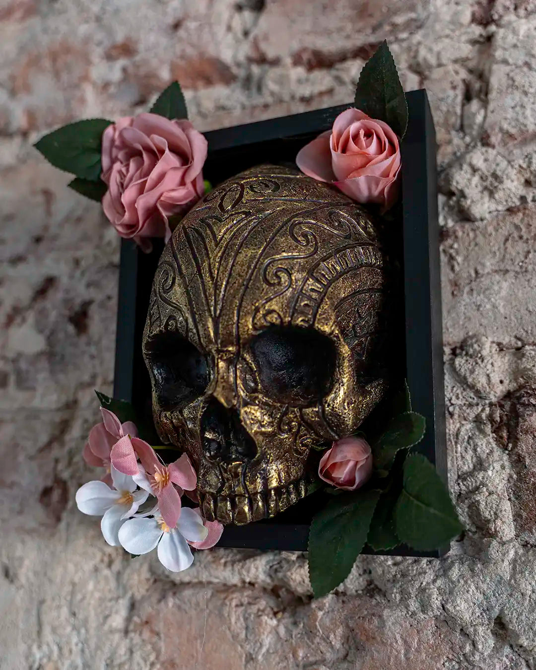 An ornate gold skull sculpture with intricate scrollwork, displayed inside a black rectangular frame and decorated with pink and white flowers on a rustic stone wall background.