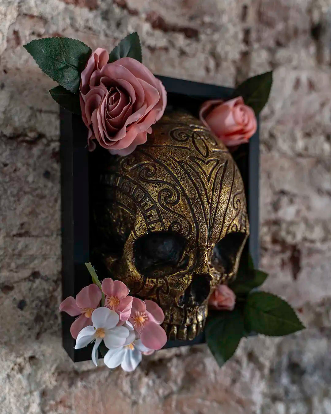 A decorative gold skull with intricate scrollwork, mounted in a black rectangular frame and adorned with pink and white artificial flowers and green leaves, set against a rustic stone wall.