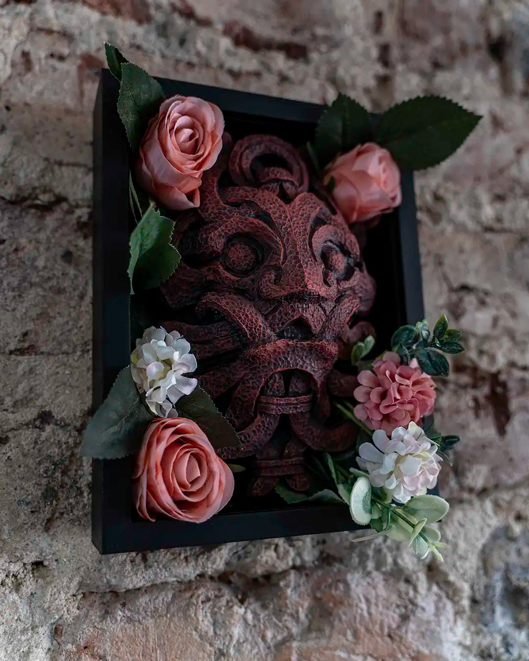 A rectangular black shadow box plaque featuring a carved dark brown tribal mask, decorated with pink and white artificial flowers and green leaves, mounted on a rustic stone wall.