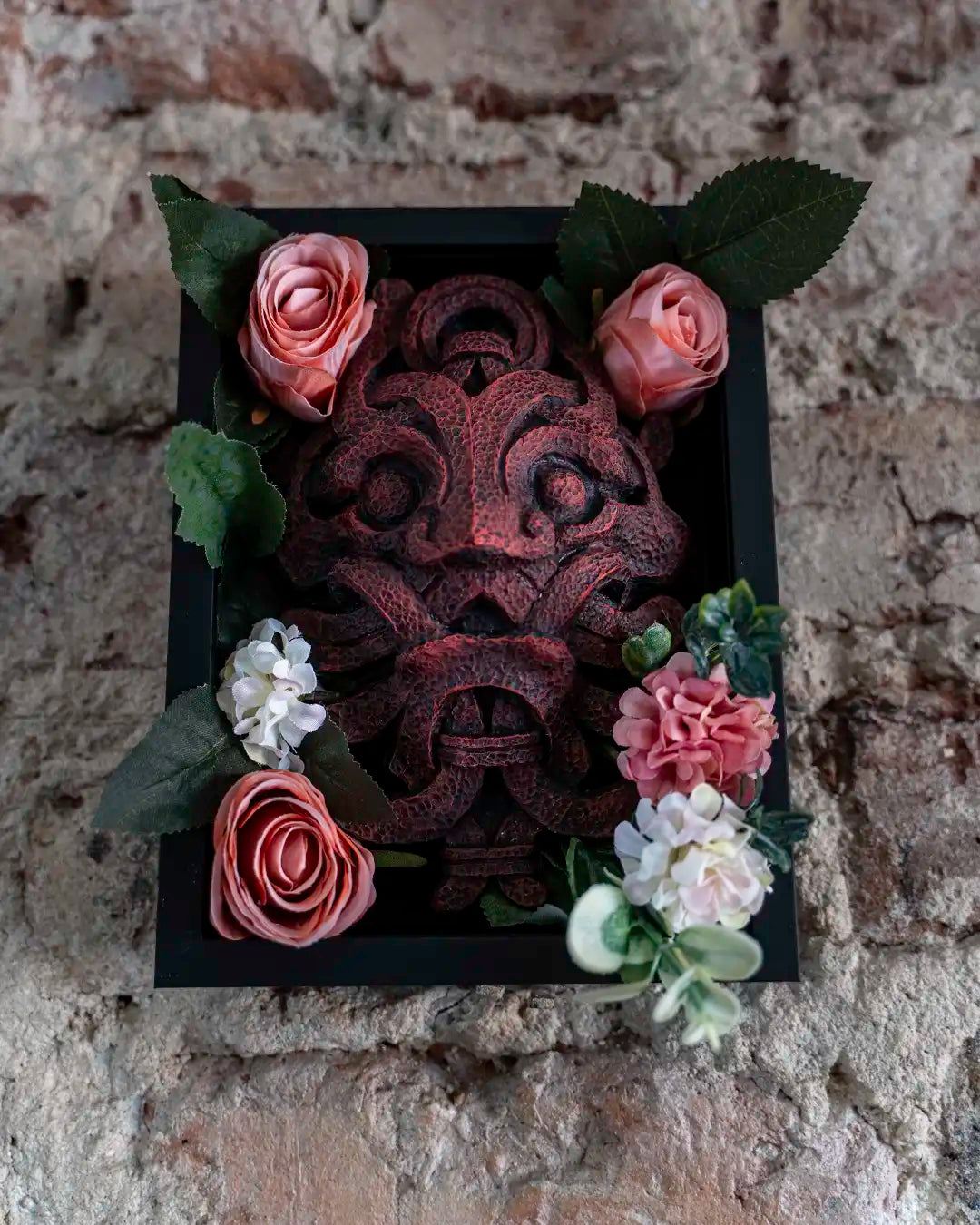 A decorative black square wall plaque featuring a dark reddish-brown carved mask, surrounded by pink and white artificial flowers and green leaves, set against a rustic brick wall background.