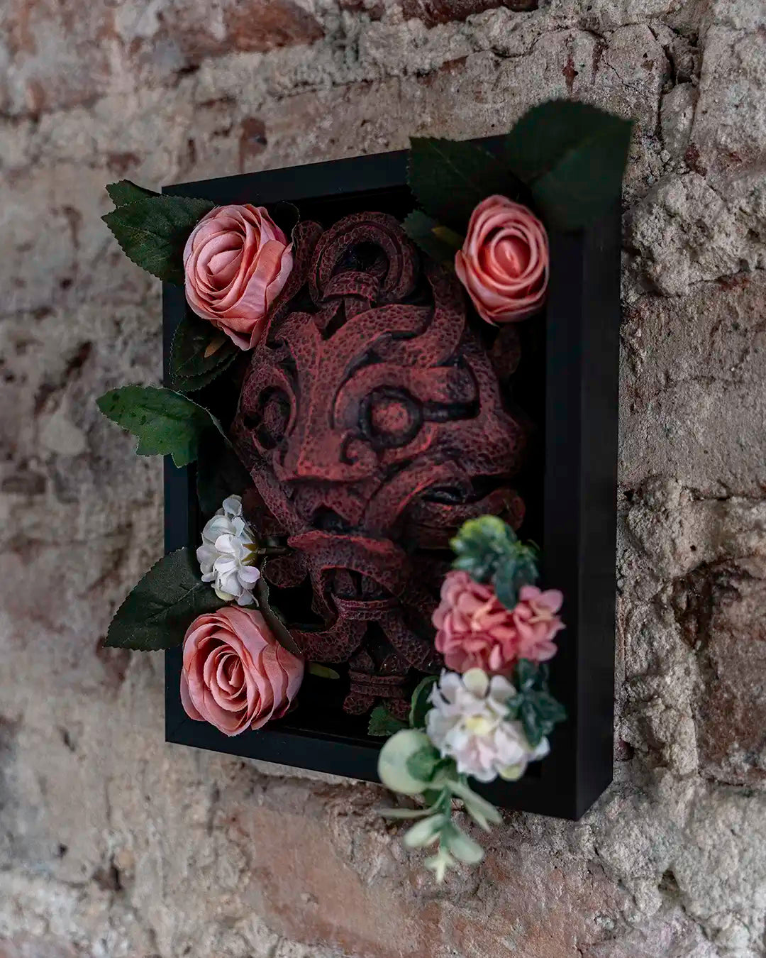 A black shadow box wall art piece featuring a carved reddish-brown mask, decorated with pink and white artificial flowers and green leaves, mounted on a rustic brick wall.