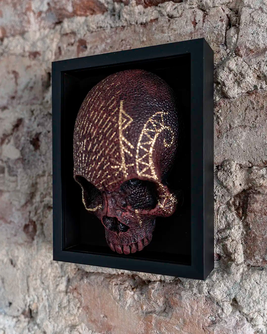 A decorative skull mounted inside a black shadow box frame, displayed against a textured stone wall. The skull has a dark reddish-brown finish with metallic gold runic symbols carved or painted across its surface.