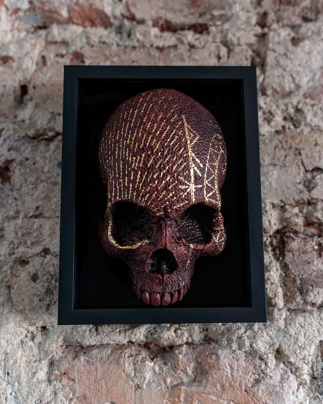 A decorative skull mounted inside a black shadow box frame, displayed against a rustic brick wall. The skull has a dark reddish-brown textured finish with gold painted geometric patterns.
