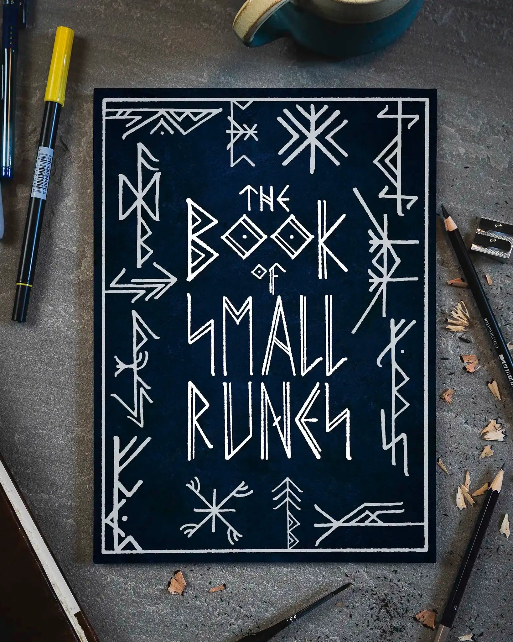 The Book of Small Runes with Norse-inspired designs, surrounded by pens, pencils, and a mug on a gray surface.