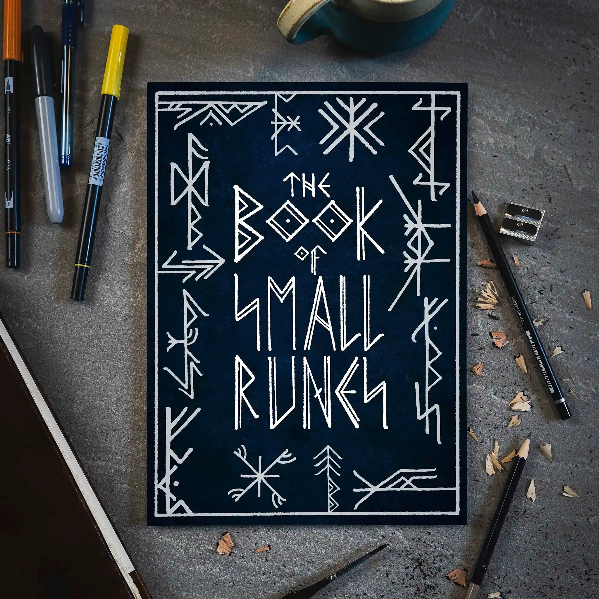 The Book of Small Runes with Norse-inspired designs, surrounded by pens, pencils, and a mug on a gray surface.