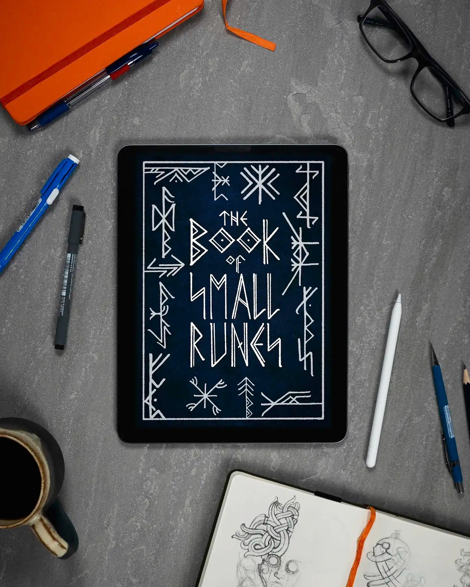 The Book of Small Runes displayed on tablet with Norse art sketches, pens, and coffee mug on desk