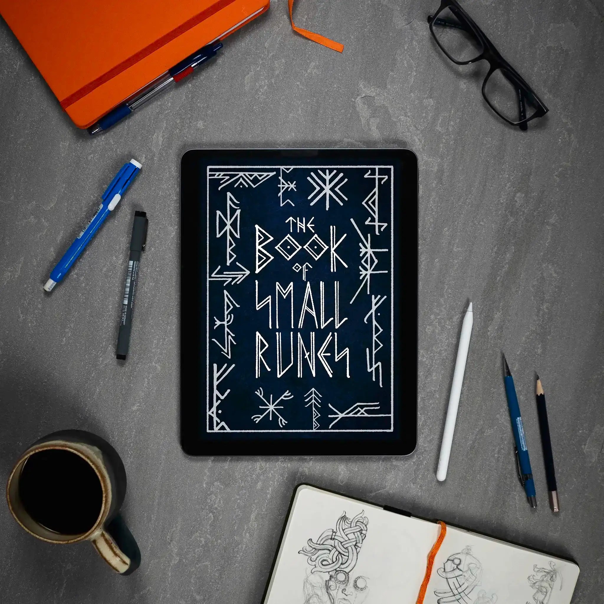 The Book of Small Runes displayed on tablet with Norse art sketches, pens, and coffee mug on desk