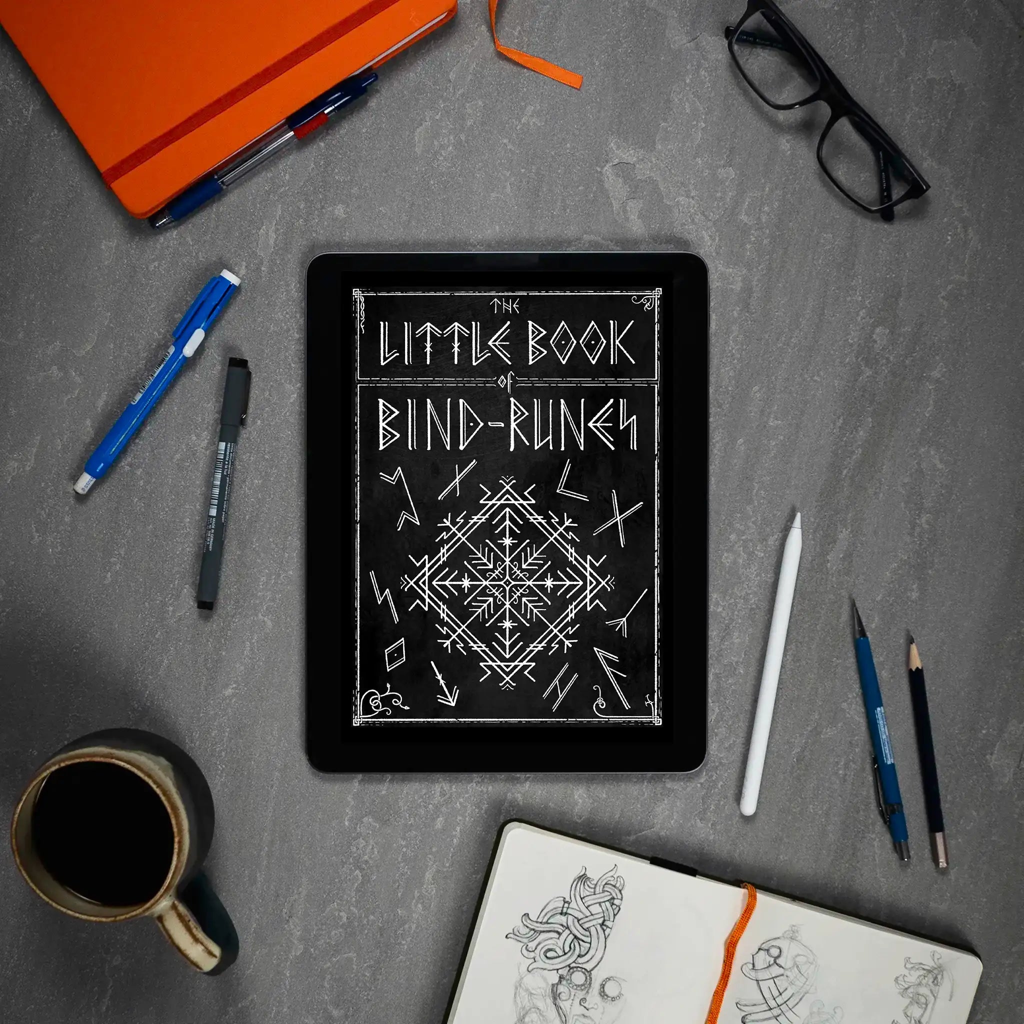 The Little Book of Bind Runes displayed on tablet with sketchbook, pens, and coffee — Norse tattoo design resource