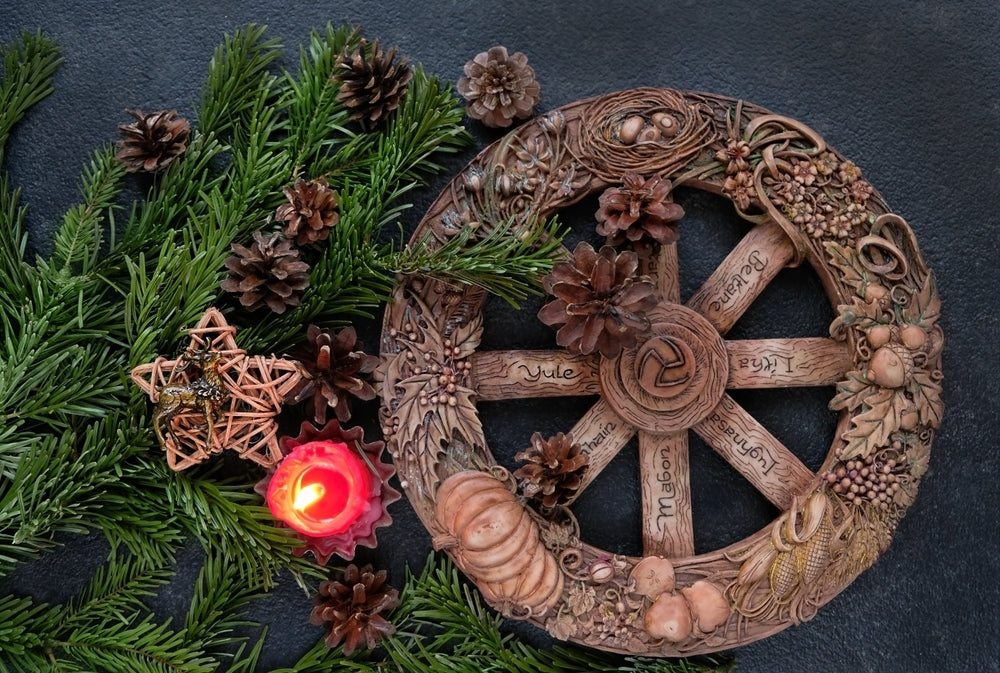 Wheel of the Year with carved seasonal names and natural decorations, surrounded by pinecones, greenery, a red candle, and a wicker star ornament on a dark background