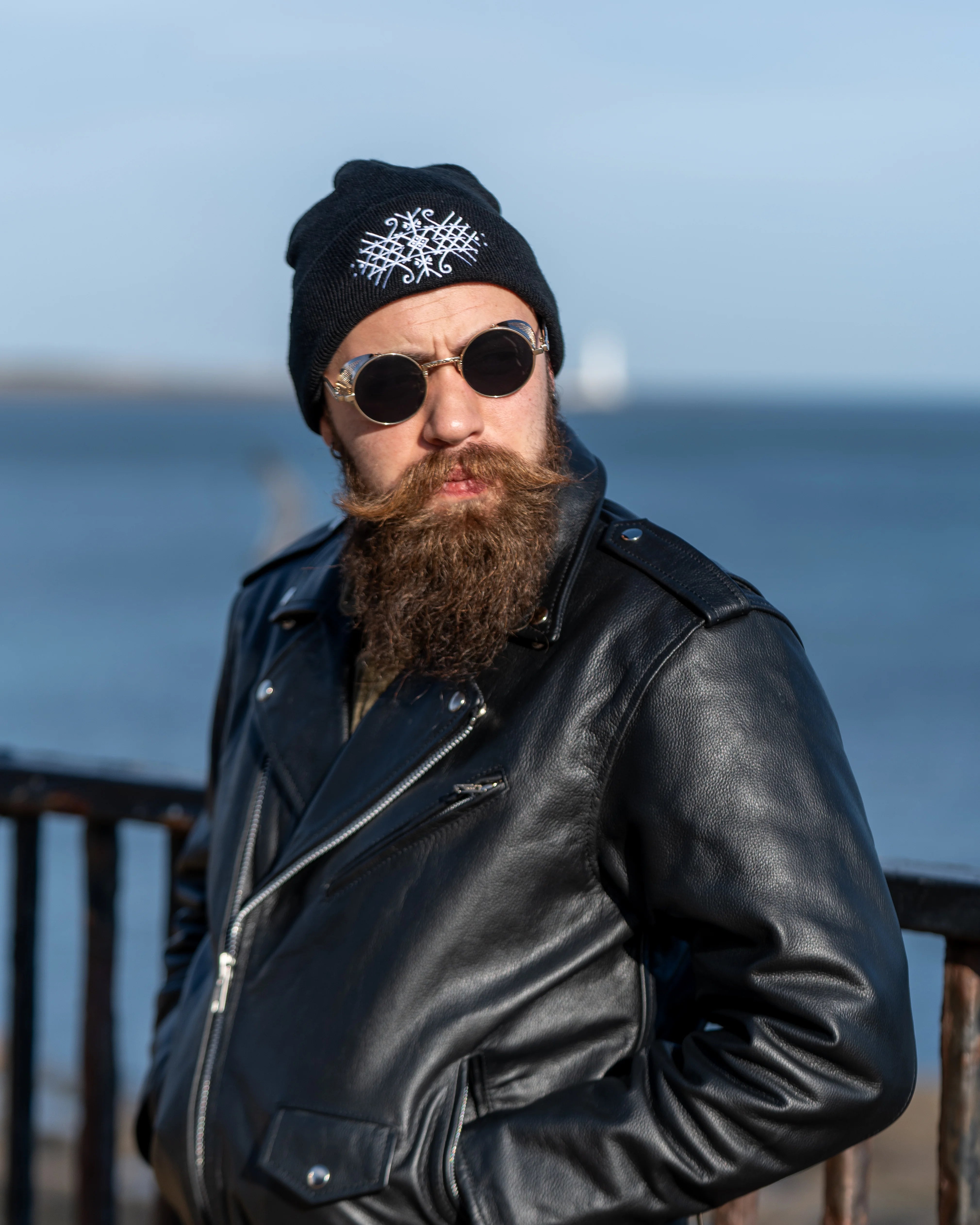 A portrait of a man with a thick brown beard wearing a black knit beanie with a white embroidered bindrune design and a black leather motorcycle jacket, set against a soft blue sky and water background.