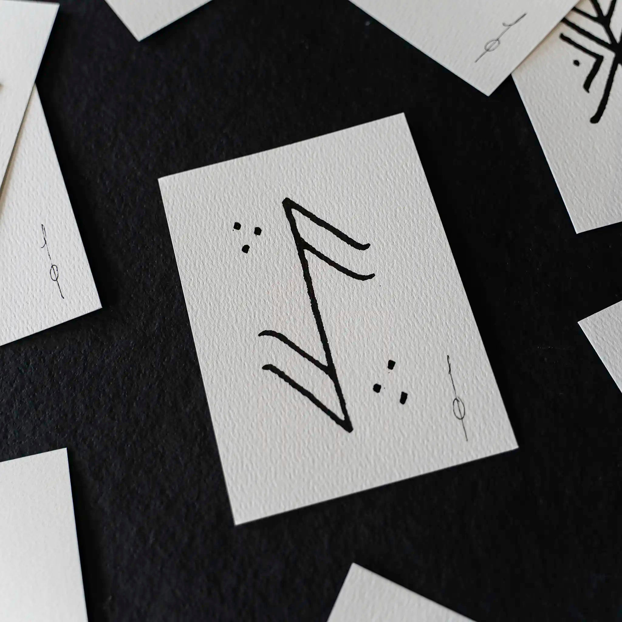 A close-up of a white textured card featuring a black hand-drawn rune symbol, surrounded by scattered matching white cards on a dark background.