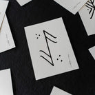 A close-up of a white textured card featuring a black hand-drawn rune symbol, surrounded by scattered matching white cards on a dark background.