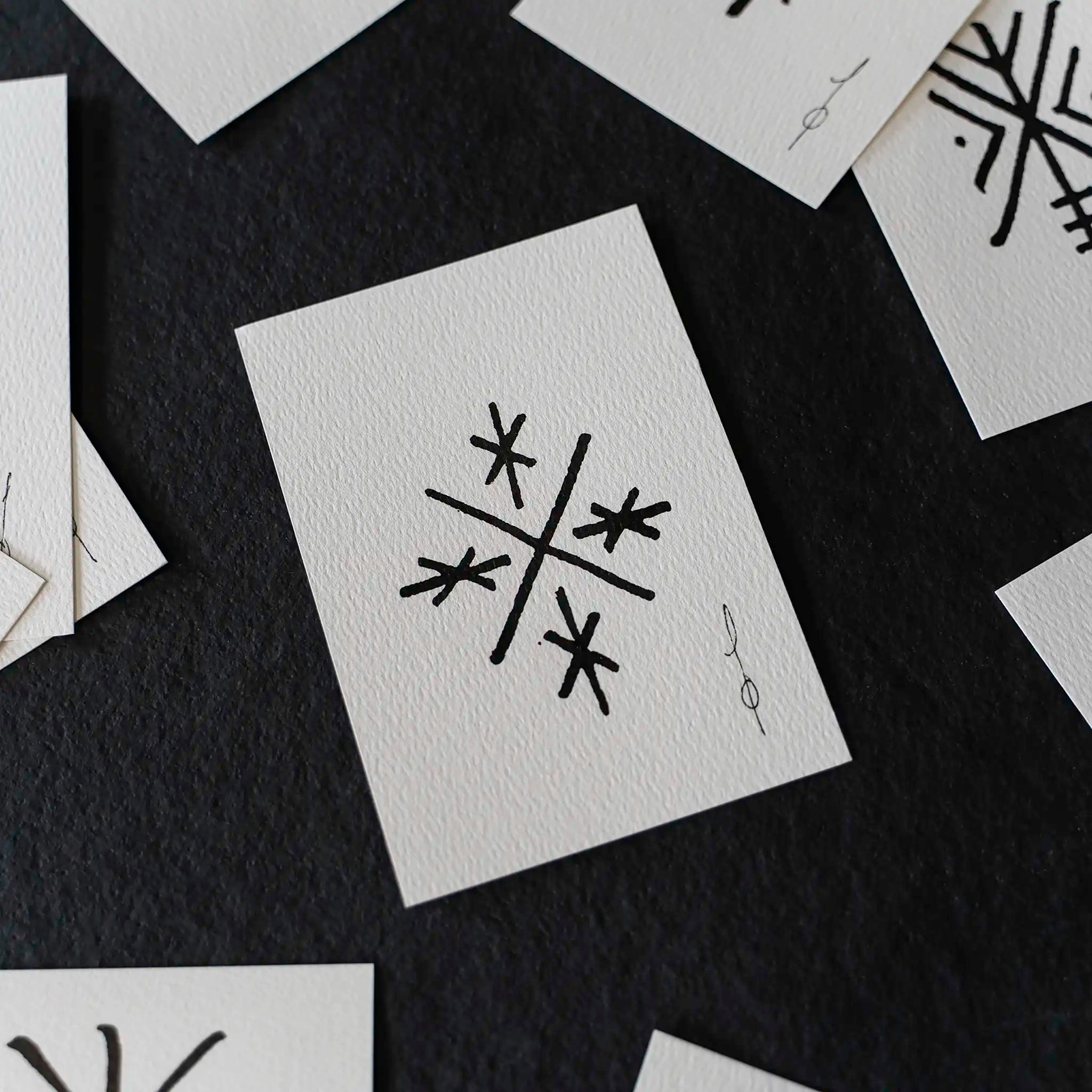 A close-up flat-lay of several white textured cards scattered on a dark charcoal background. The central card shows a black abstract geometric symbol resembling a stylized snowflake or starburst, with a small vertical signature-like mark beside it.