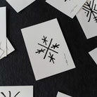 A close-up flat-lay of several white textured cards scattered on a dark charcoal background. The central card shows a black abstract geometric symbol resembling a stylized snowflake or starburst, with a small vertical signature-like mark beside it.