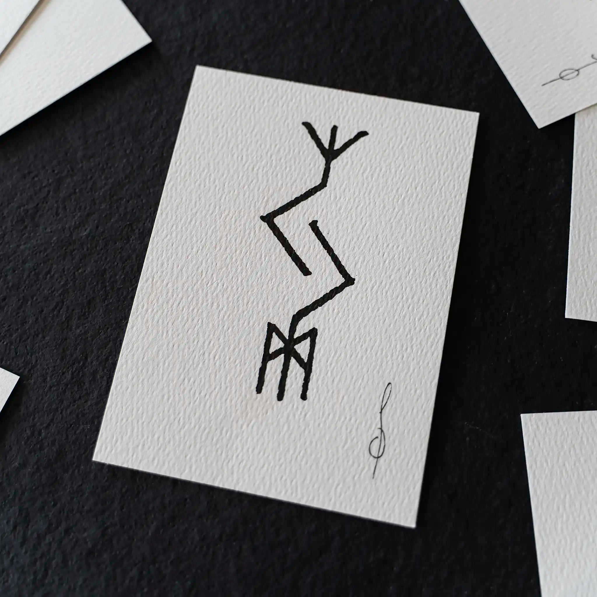 A square white art print with a black abstract line drawing, displayed on a dark textured surface with other partial prints around it.