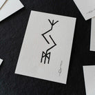 A square white art print with a black abstract line drawing, displayed on a dark textured surface with other partial prints around it.