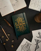 Dark green notebook with gold Norse rune design, surrounded by runestones, sketches, and Viking art tools